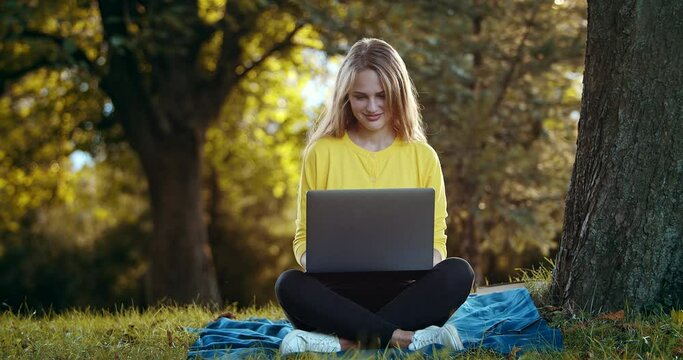 Front view of happy female in yellow sweater and black trousers typing on portable laptop during sunset at summer park. Concept of people, technology and nature.
