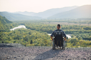 Lonely young man in a wheelchair enjoying fresh air in sunny day on the mountain. People with disabilities travelling.