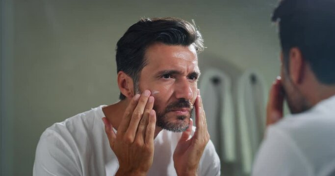 An young smiling man is applying after cleaning his face day or night cream to take care of skin behind a mirror in a bathroom.