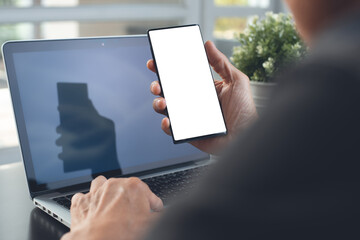 Mockup of man hand using blank screen mobile phone and working on laptop computer in office