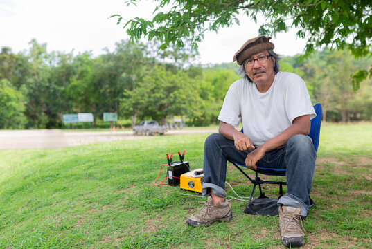 Smart Old Asian Man Sitting On Seat Near Battery Charger, He Camping In Forest, Happiness Retirement Activity