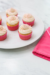 Close Up of Vanilla Cupcakes with Vanilla Icing in Pink Paper Cups with Pink Sprinkles on a White Plate on White Countertop with Pink Napkin Beside
