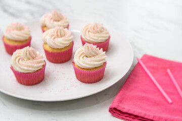 Close Up of Vanilla Cupcakes with Vanilla Icing in Pink Paper Cups with Pink Sprinkles on a White Plate on White Countertop with Pink Napkin Beside