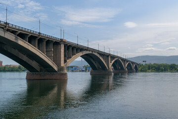 Naklejka na ściany i meble Old concrete bridge over a large river. Green trees on the shore, mountains. The background is the sky. Communal bridge in the city of Krasnoyarsk across the Yenisei River.