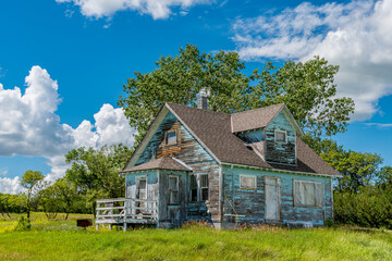Old, abandoned blue prairie farmhouse with trees, grass and blue sky in Kayville, Saskatchewan,...