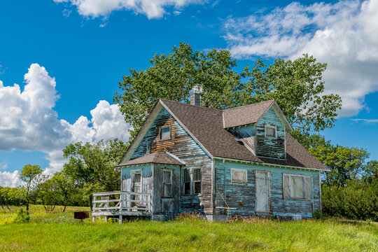 Old, abandoned blue prairie farmhouse with trees, grass and blue sky in Kayville, Saskatchewan, Canada