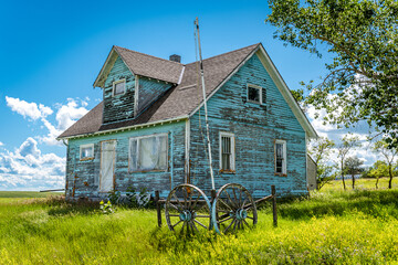 Old, abandoned blue prairie farmhouse with trees, grass, blue sky and a wagon wheel in Kayville,...