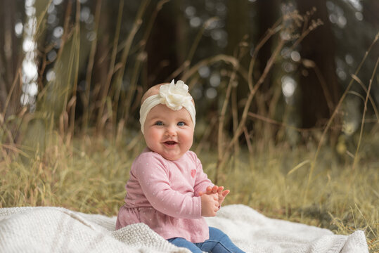 Adorable baby in pink shirt with white head band sitting smiling on blanket in park