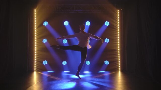 Professional male ballet dancer performs a pirouette in a dark smoky studio. Silhouette of men against the background of blue lights in slow motion.
