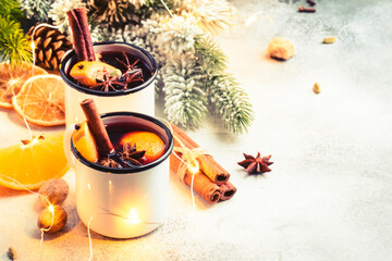 Fototapeta na wymiar Mulled wine in white metal mugs with cinnamon, spices and orange with fir tree and christmas lights - traditional drink on winter holiday. Copy space, white background
