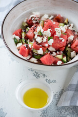Bowl of salad with watermelon cubes, feta cheese and cucumber slices, vertical shot on a white concrete surface, close-up