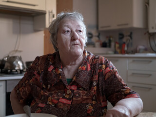 Funny old woman with gray hair and wrinkled face. pensioner suffers from senile memory loss and is upset because of her dependence on relatives and carers. Woman sitting at table in kitchen