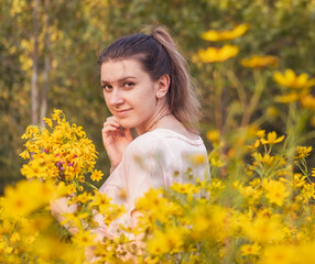 Naklejka premium Environmental portrait of young attractive woman looking back while standing in field of tall yellow wildflowers at sunset in early fall