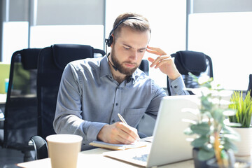 Smiling male call-center operator with headphones sitting at modern office, consulting online...