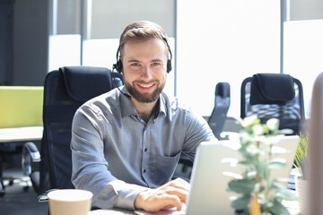 Smiling male call-center operator with headphones sitting at modern office, consulting online...