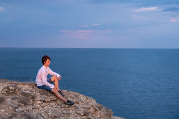 Naklejka na ściany i meble A young man sits on a rocky seashore at sunset.