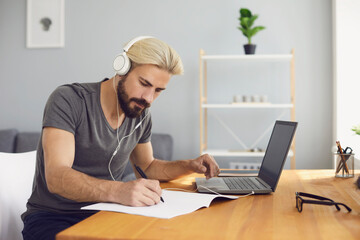 Online education. A young man sitting at a table at home using a laptop and a video call to listen to a lesson online.