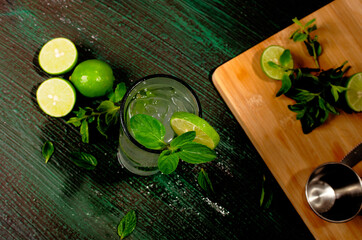 Mojito, cóctel de origen cubano, sobre mesa verde decorada con ingredientes del cóctel. Fondo de madera.