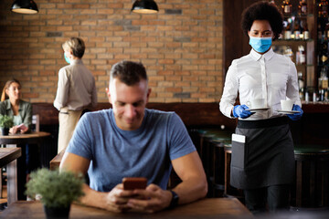 Black waitress wearing face mask and gloves while serving coffee in a cafe during coronavirus epidemic.