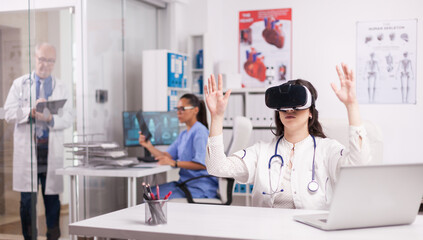 Young medic doing hand gesture wearing virtual reality headset in hospital office. Doctor in white coat with stethoscop. Senior physician writing notes on clipboard. Nurse in blue uniform looking at x