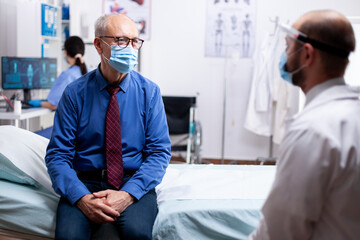 Obraz premium Sick patient talking with doctor during consultation wearing protective mask against covid in hospital consultation room. Global health crisis, medical system during pandemic, sick elderly patient.