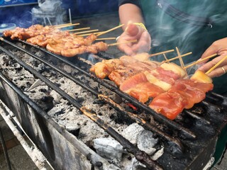 Closeup pork with bamboo stick grilled on the charcoal grill with smoke and motion blurred hands of seller in background. Famous street food in morning.