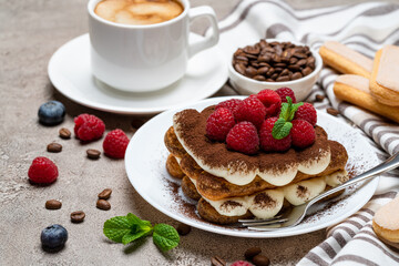 portion of Classic tiramisu dessert with raspberries and cup of coffee on grey concrete background