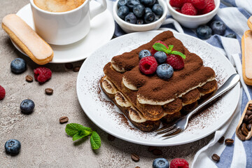 portion of Classic tiramisu dessert with raspberries, bluebedrries and cup of coffee on grey concrete background