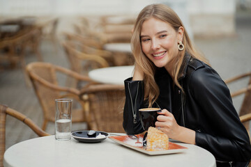 a beautiful young woman of Caucasian appearance is sitting in a cafe and smiling. Summer Breakfast outside. Earrings in the ears, stylish clothes.