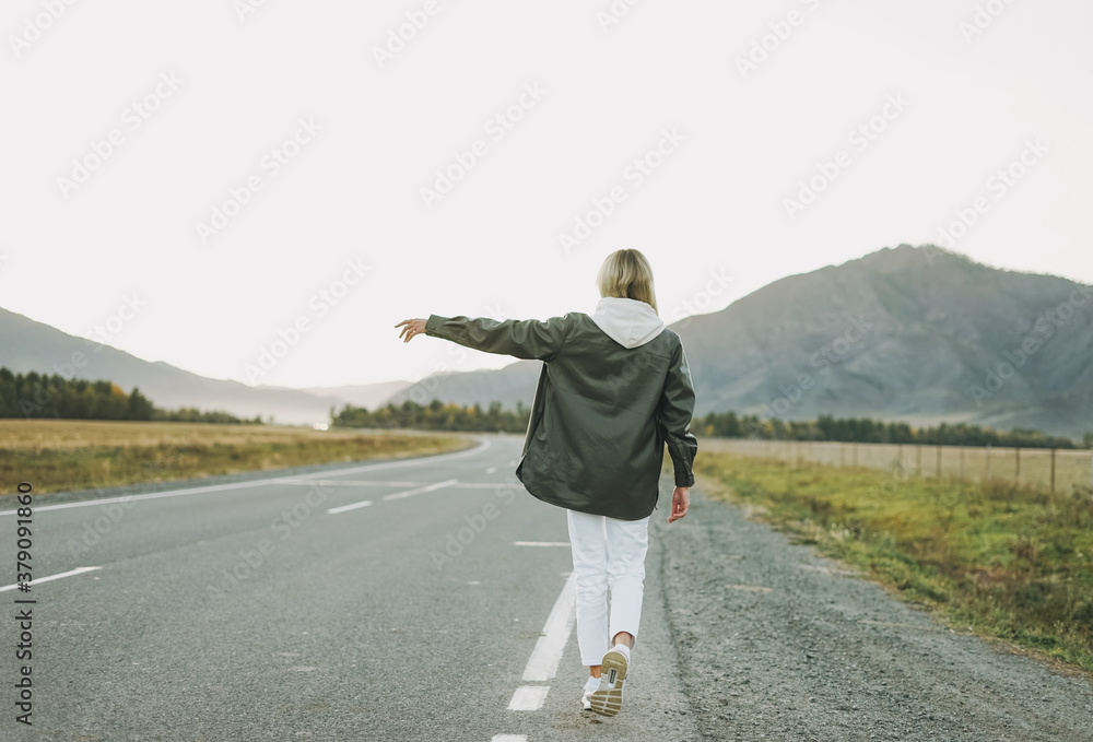 Sticker Blonde young woman traveler in white hoodie on road, trip to mountains, hitching a ride - Stickers