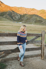 Young blonde woman in nordic sweater and jeans near old wooden fence on the mountains background