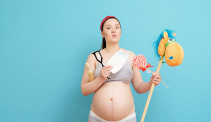 surprised young pregnant woman on blue background holding toys, diaper, cooking appliances in her hands