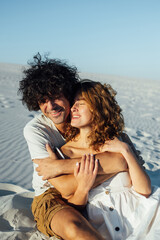 a guy with curly hair and his girlfriend are hugging cheerfully on the sand