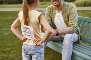 Naklejka premium Father sitting on the wooden bench in park while his little daughter hiding handmade postcard for him, small girl making a surprise for Happy Fathers Day