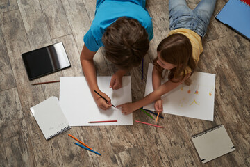Top view of little caucasian boy and girl lying on the wooden floor at home and drawing on a white sheet of paper with colorful pencils