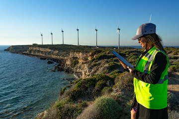 Female engineer standing with digital tablet against wind turbine on sunny day, looking at wind...