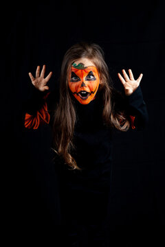 girl with pumpkin makeup scares and pretends to be a monster on a black background, Halloween celebration, low key.