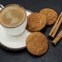 Cup with hot coffee and saucer on a black background. There are small bubbles on the surface of the drink. Next to it are three cookies and two cinnamon sticks. The dishes are white with beige dots.