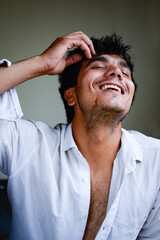 indoor portrait shot of a young handsome Indian man wearing a white unbuttoned shirt and laughing. he has a hairy chest. one hand is lifted