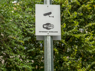 A sign on a metal pole in a park with Wi Fi internet wireless and a security camera