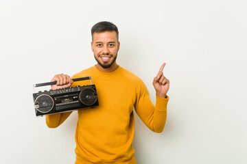 Young south-asian man holding a guetto blaster smiling cheerfully pointing with forefinger away.