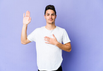 Young caucasian man isolated on purple background taking an oath, putting hand on chest.