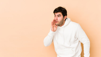 Young man isolated on beige background is saying a secret hot braking news and looking aside