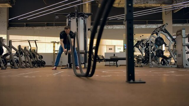 Powerful caucasian athlete trains with ropes in the gym. He makes the waves heavy, large rooms in the horizontal plane, modern exercise machines in the background.