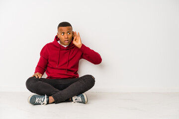 Young latin man sitting on the floor isolated trying to listening a gossip.