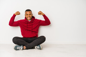 Young latin man sitting on the floor isolated showing strength gesture with arms, symbol of feminine power