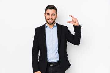 Young caucasian business man against a white background isolated holding something little with forefingers, smiling and confident.