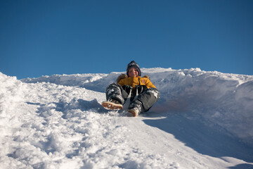 Naklejka na ściany i meble happy boy sliding down snow hill on sled outdoors in winter, cold season concept