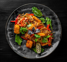 Tasty asian noodles with fried cheese tofu and vegetables in plate on black wooden table background