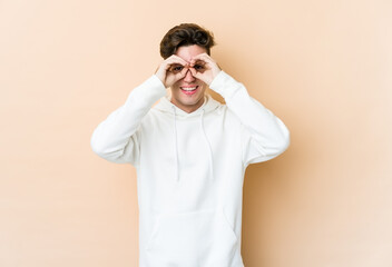 Young caucasian man isolated on beige background showing okay sign over eyes